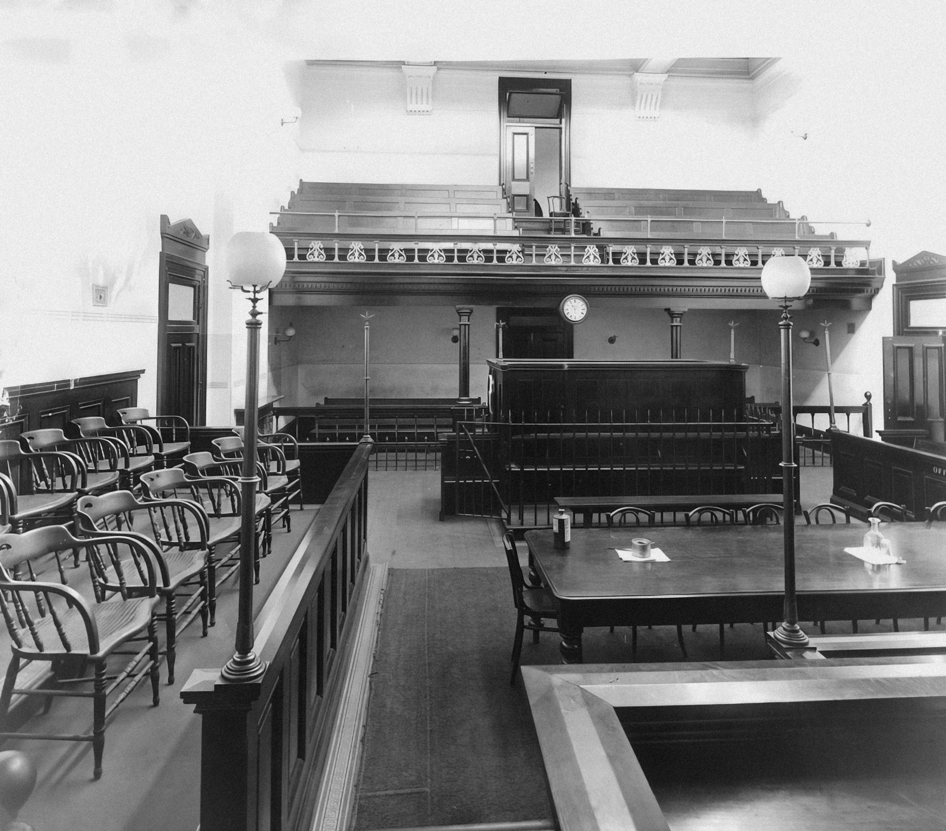 Black-and-white view of an empty historic courtroom with wooden benches, counsel tables and a raised judge's bench
