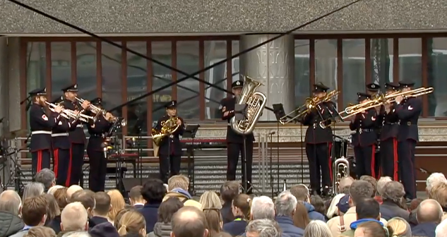 11 uniformerte musikere fra Forsvarets stabsmusikkorps på en scene foran tilskuere. De står i ujevnt fordelt halvsirkel, alle med messinginstrumenter, trompeter og horn til venstre, tubaist i midten og tromboner til høyer.