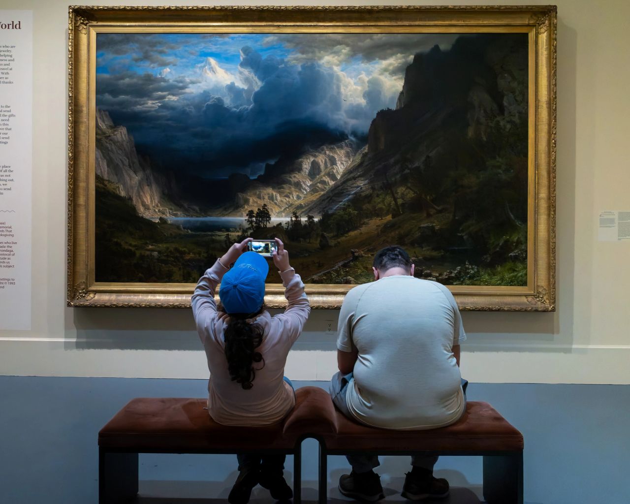 Two museum visitors sit on a bench facing a large gold-framed landscape painting; one holds a phone up to photograph the canvas.