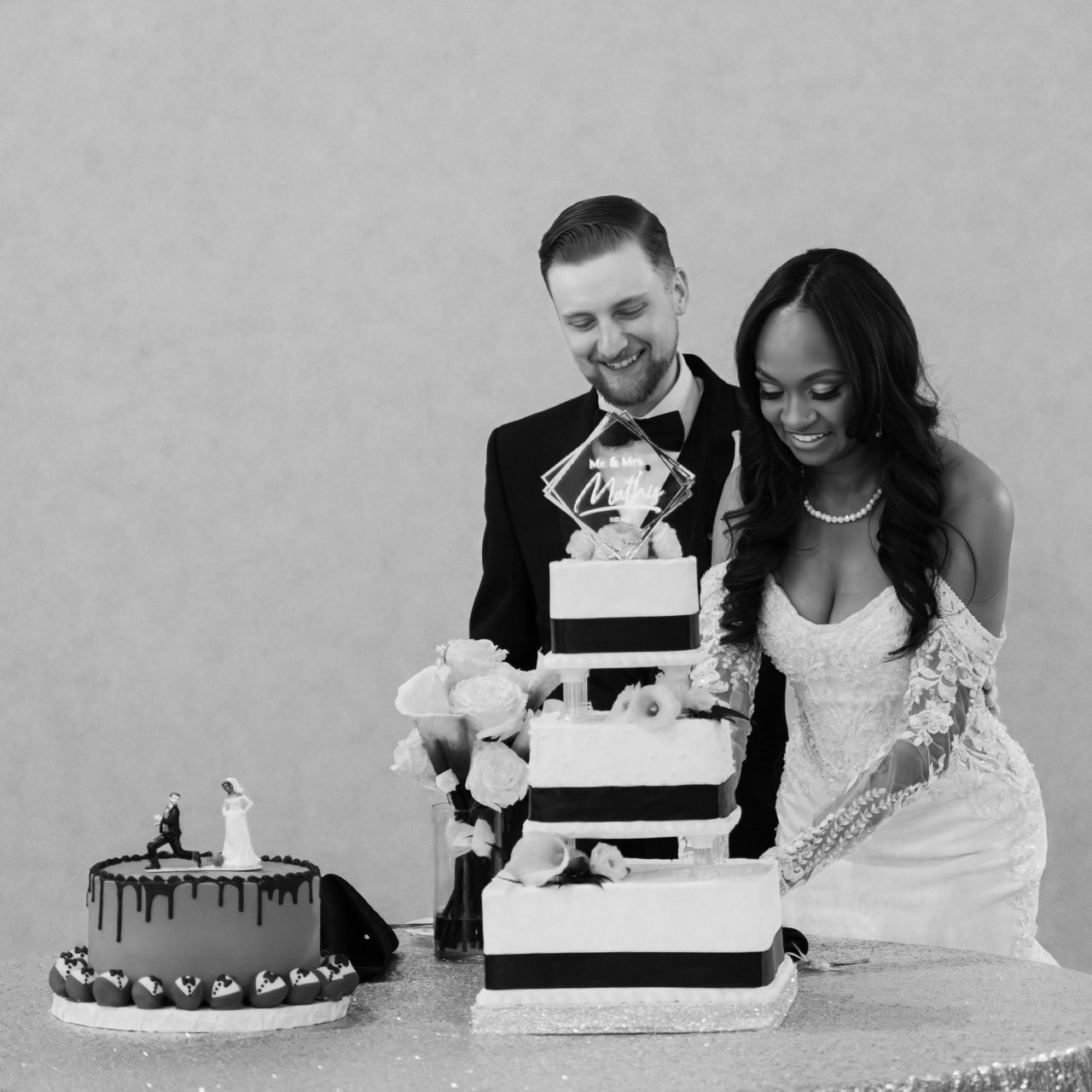 Bride in lace gown and groom in tuxedo cut a tiered wedding cake beside a smaller drip cake with figurine topper, black and white.