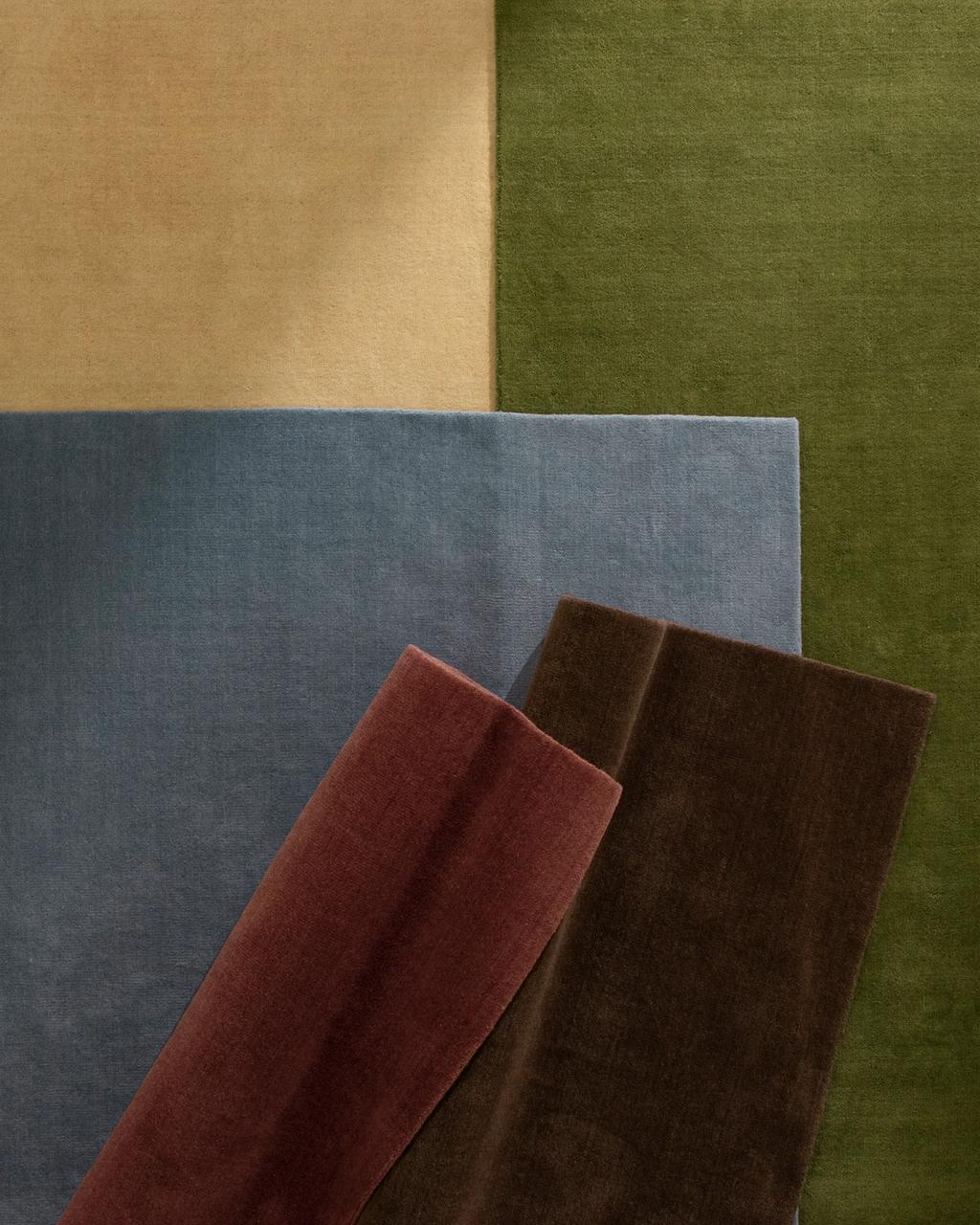 Stack of solid-color wool rugs in tan, olive green, blue, burgundy, and dark brown