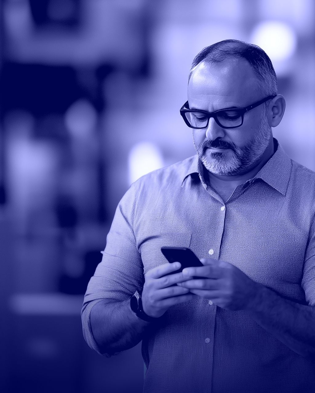 A man wearing glasses looks down at a smartphone he holds in both hands, shown in a blue-toned photo.