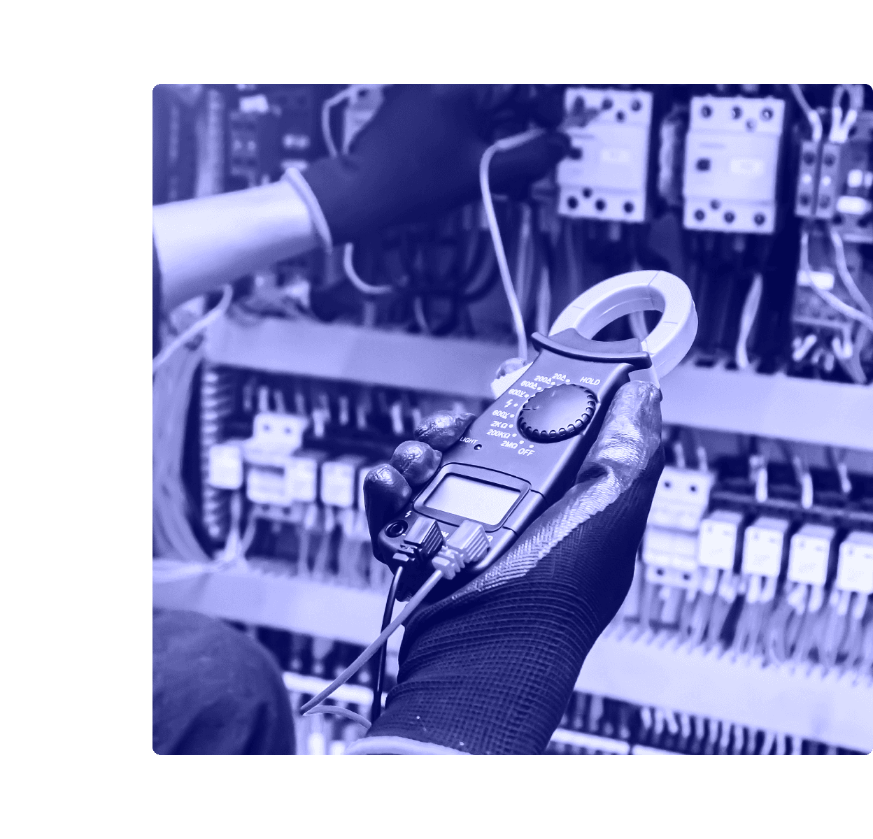 A gloved technician holds a digital clamp meter in front of an industrial electrical control panel full of breakers and wiring.