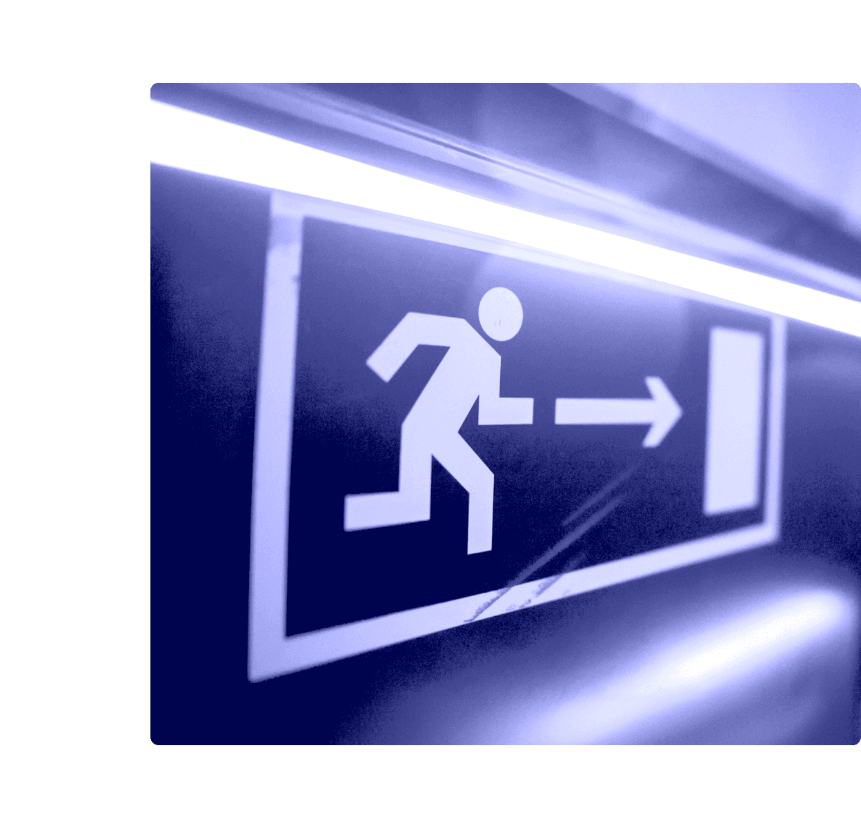 A wall-mounted emergency exit sign showing a running figure and arrow, lit by a fluorescent tube in a dim corridor.