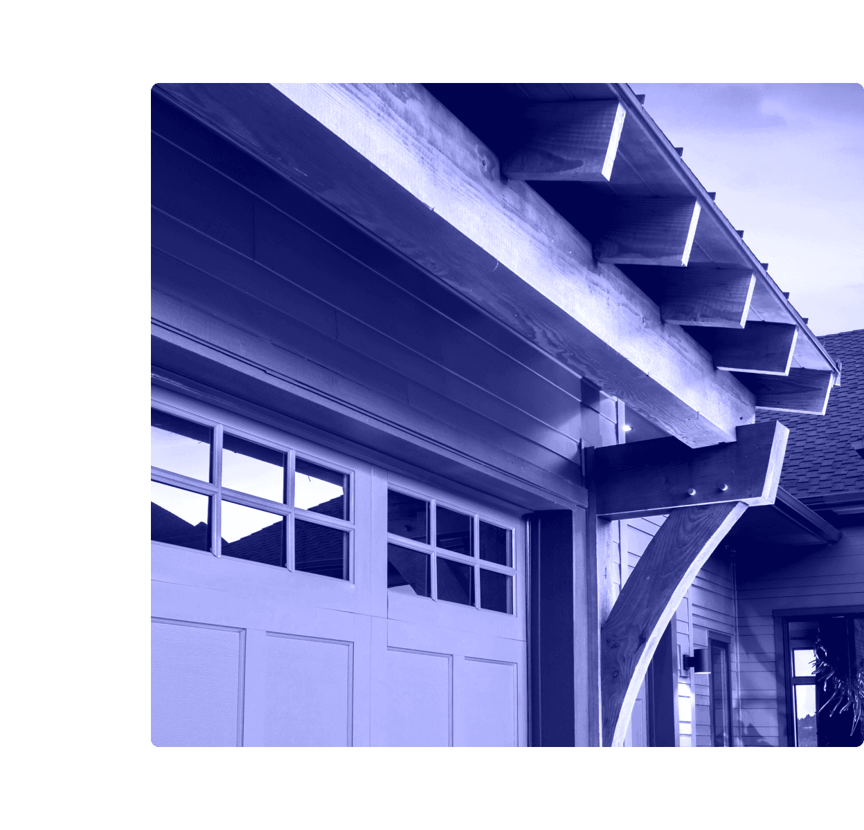 The exterior of a home showing a paneled garage door with window inserts beneath heavy wood beams and a bracketed overhang.