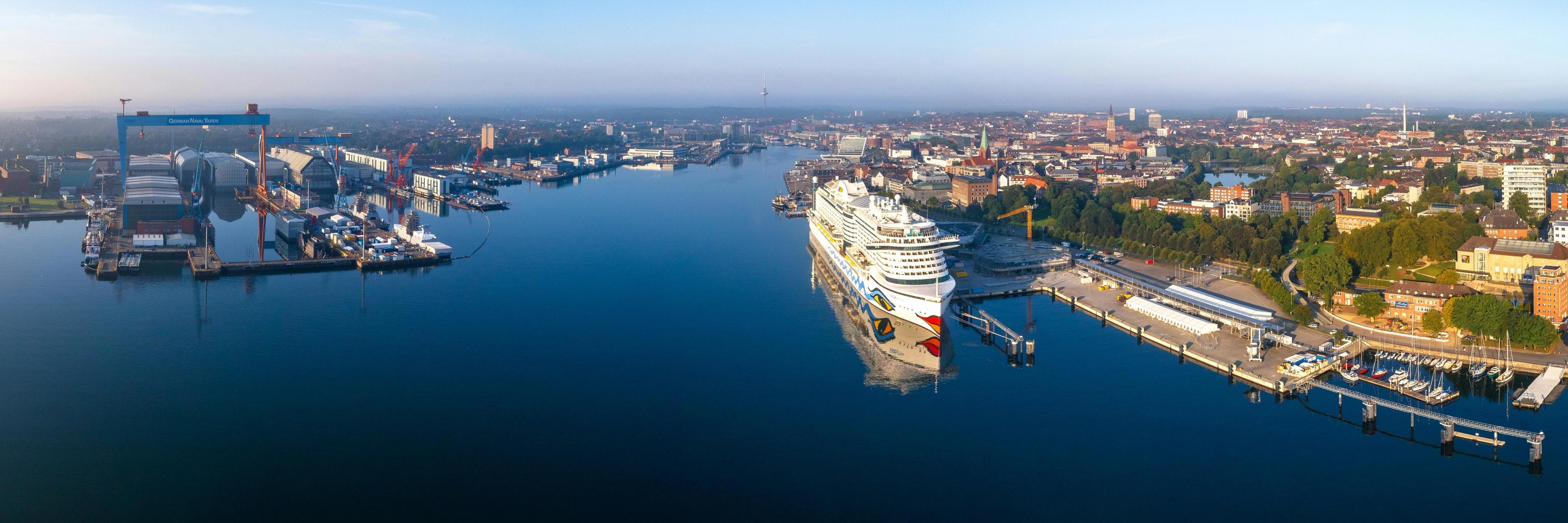 Panorama des Kieler Hafens mit Kreuzfahrtschiff am Pier, umgeben von Stadtgebäuden und Grünflächen.