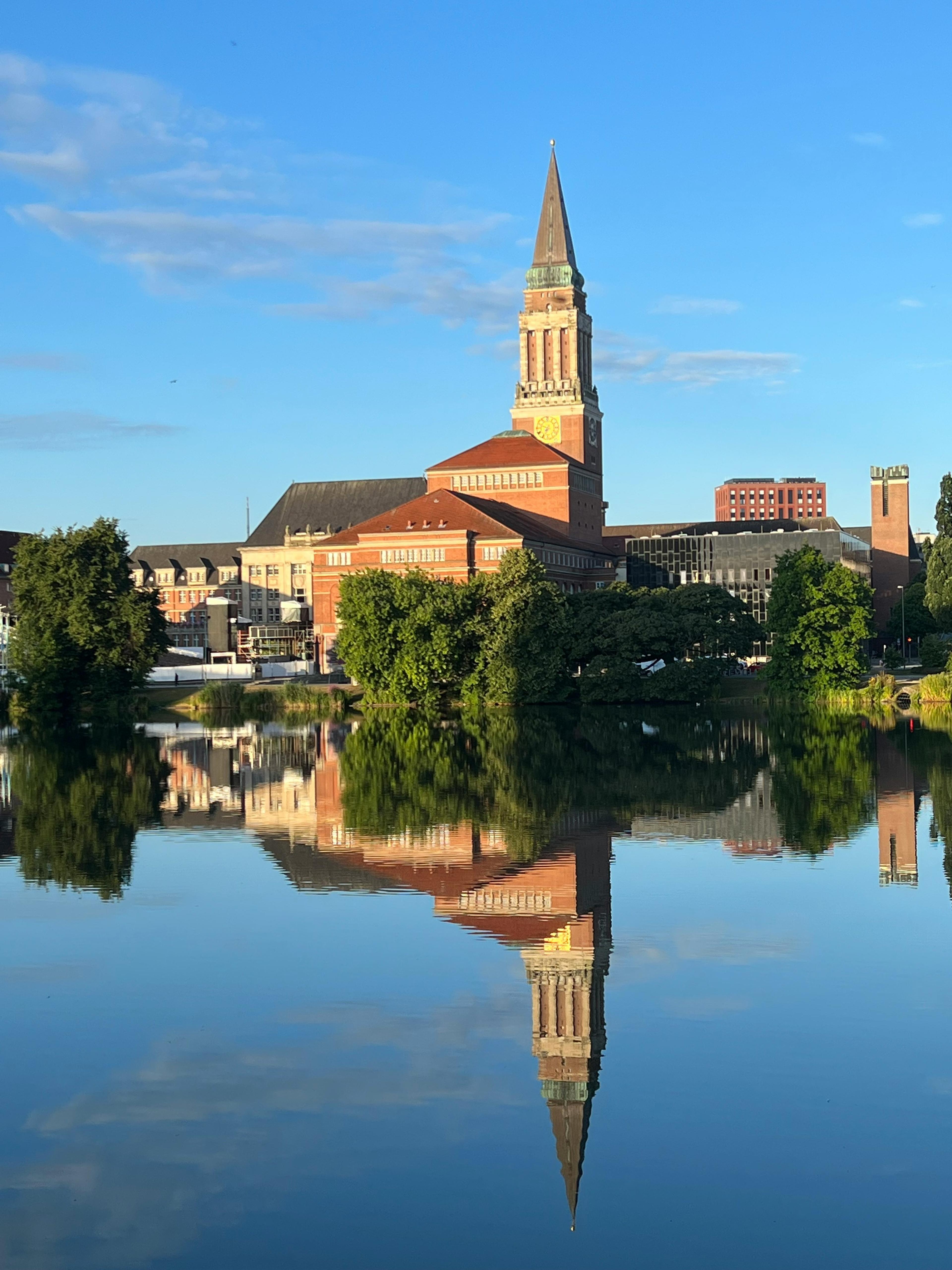 Rathaus von Kiel mit spitzem Turm, das sich im ruhigen Wasser spiegelt.