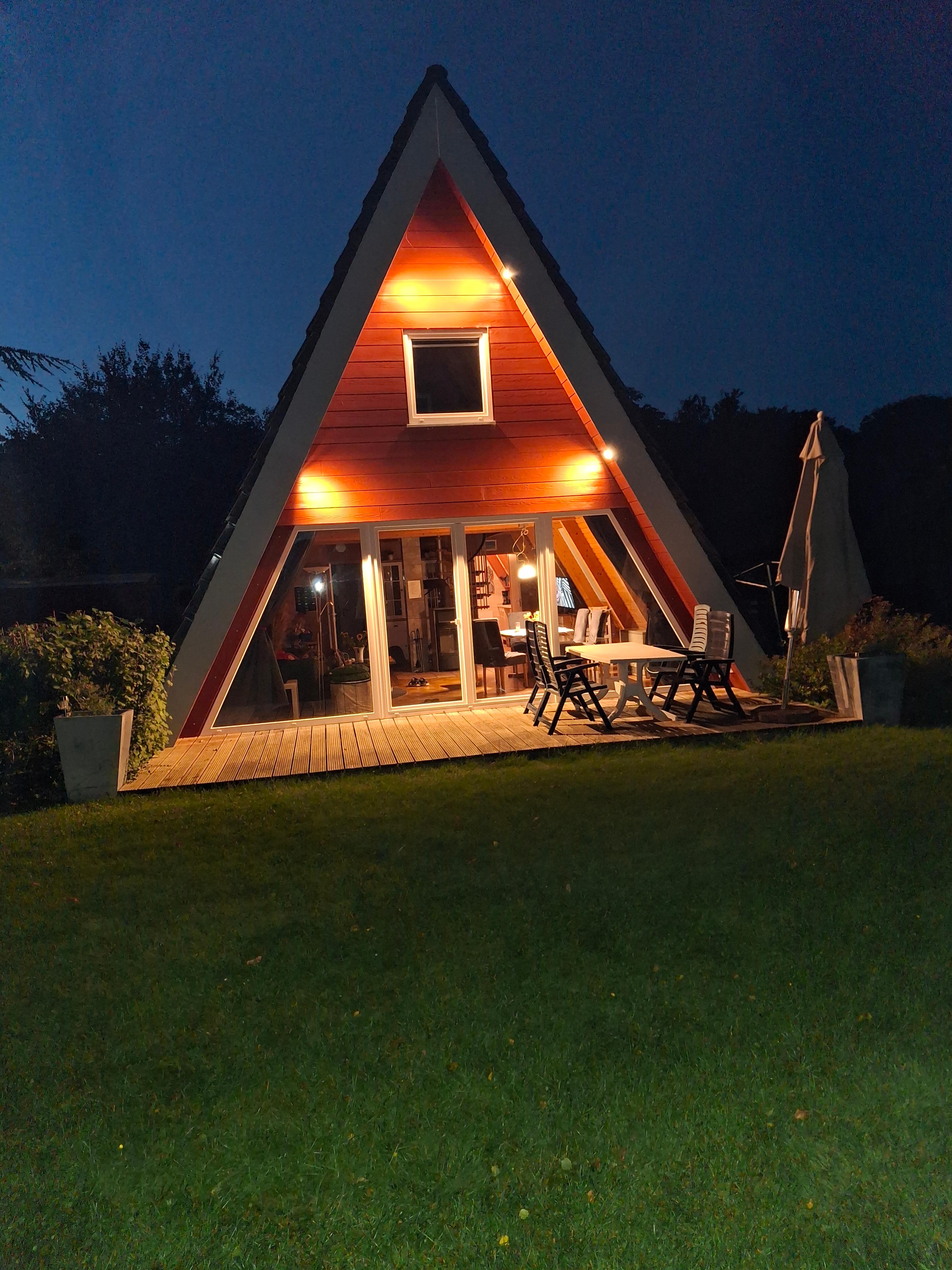 A-förmiges Holzhaus beleuchtet bei Nacht mit Terrasse und Gartenstühlen