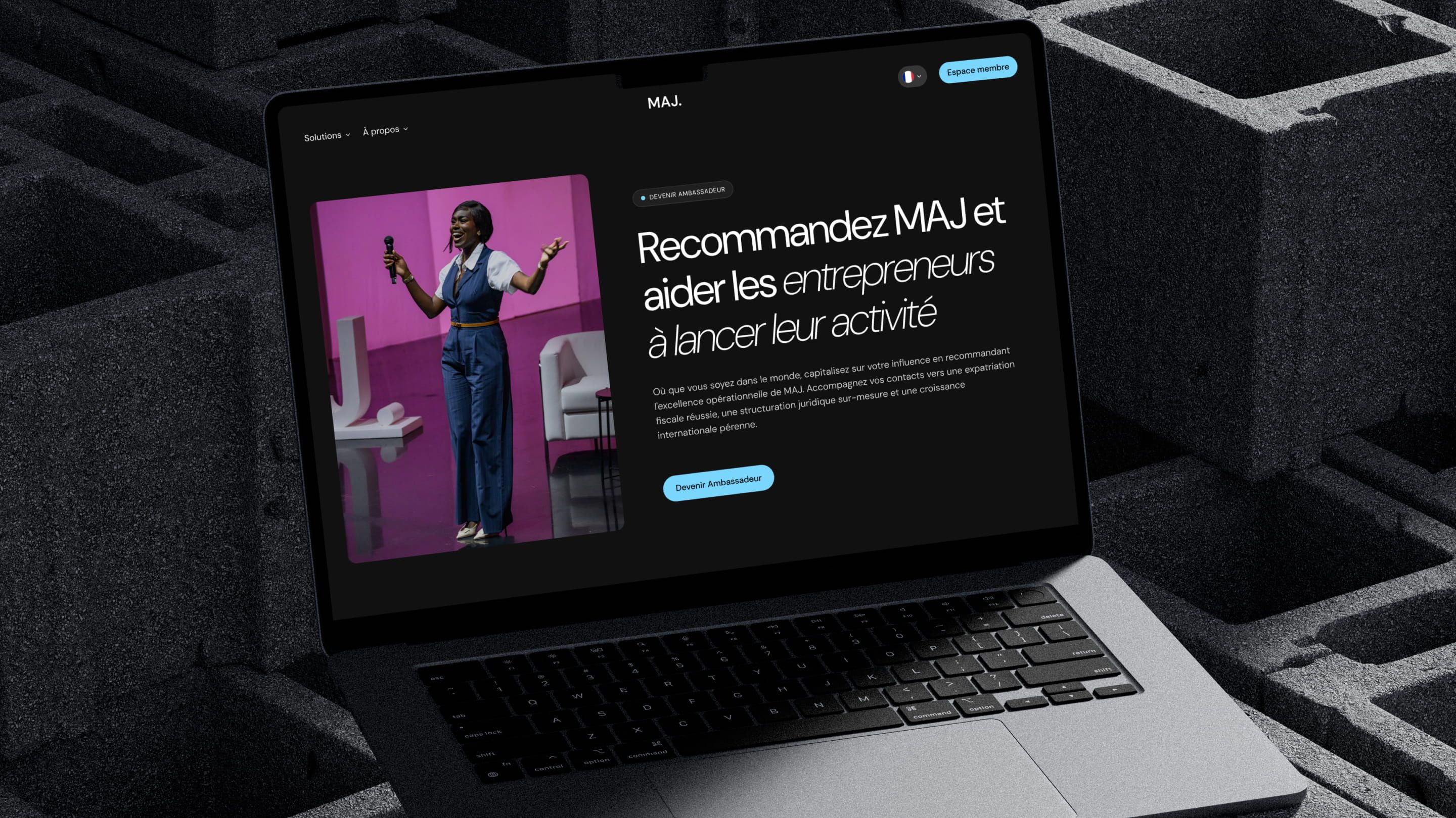 Laptop showing MAJ's dark-mode ambassador page: “Recommandez MAJ”, woman with microphone on a pink stage
