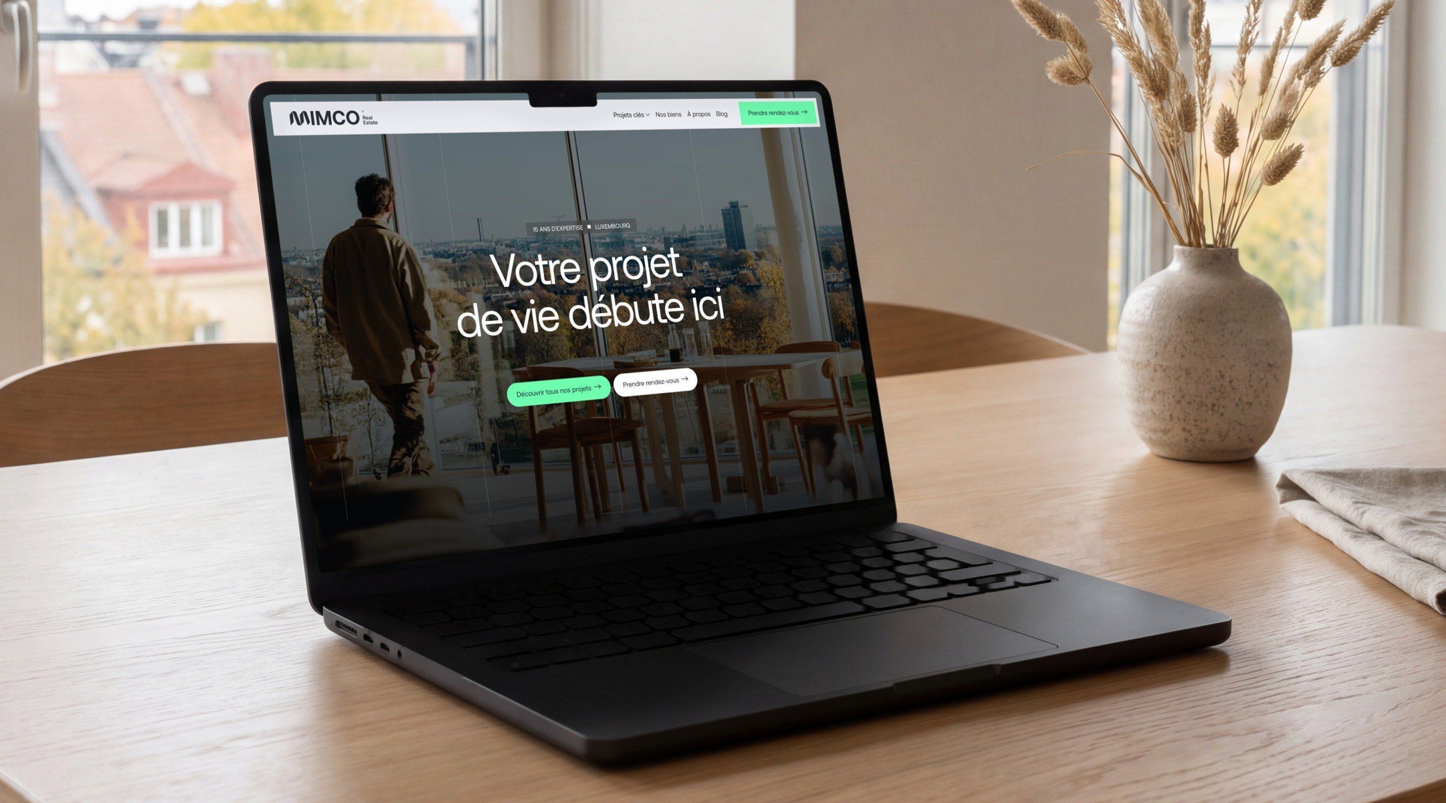 MacBook Pro on an oak table in a bright flat, open on the Mimco home page, “Your life project starts here”