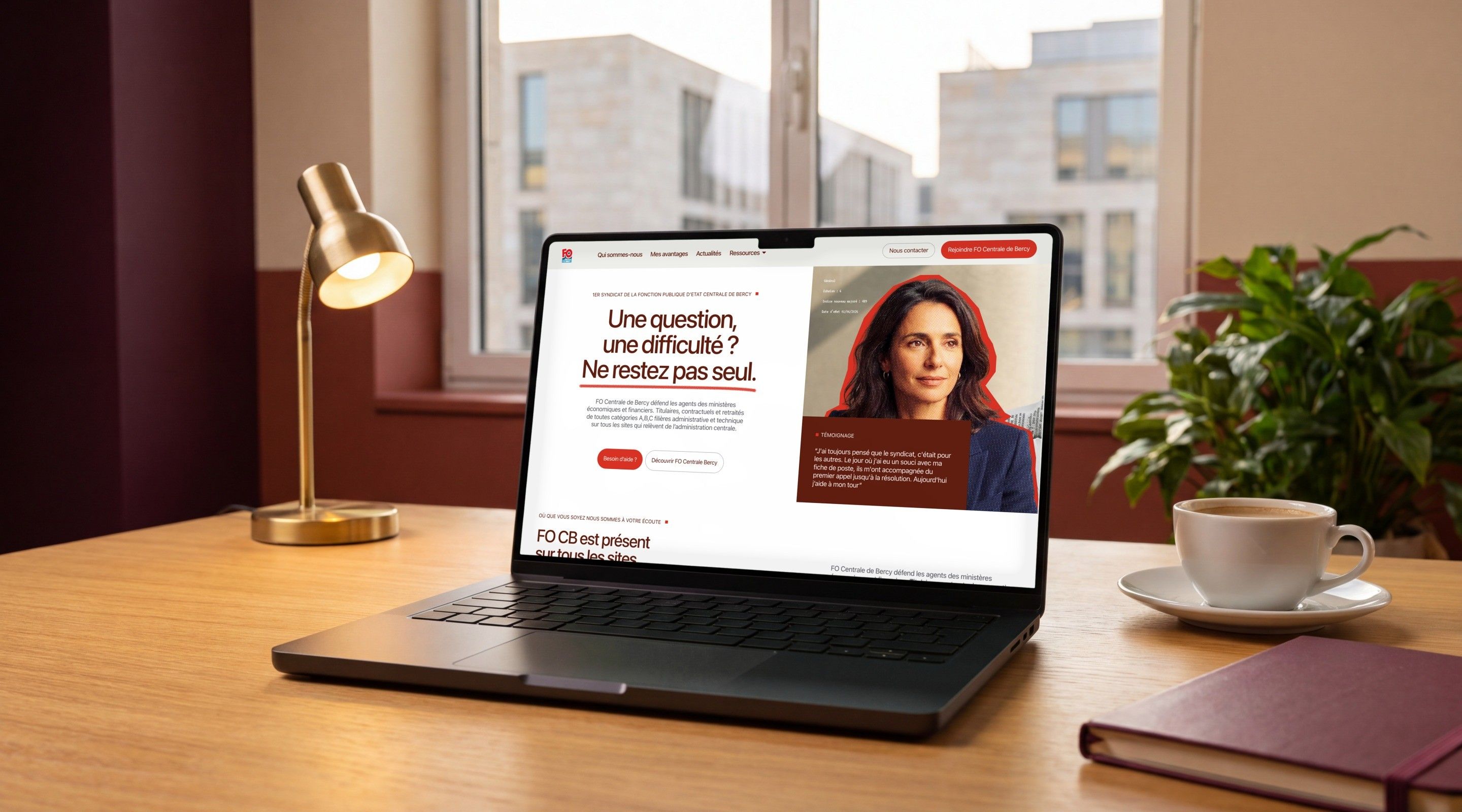 MacBook Pro open on the FO Centrale Bercy home page, “A question, a difficulty? Don't stay alone.”, on a wooden desk next to a lamp and a cup of coffee
