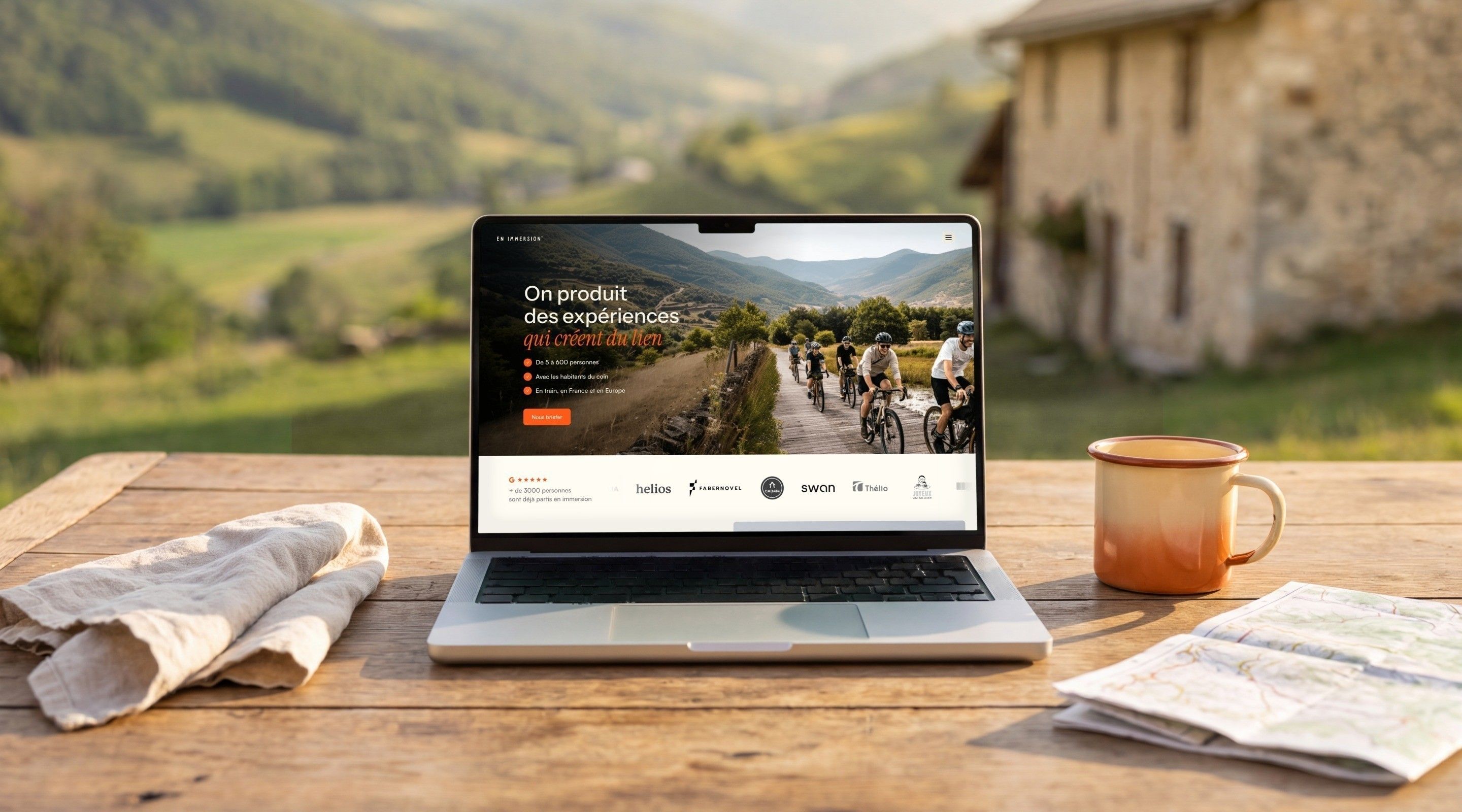 MacBook Pro open on the En Immersion home page, “We create experiences that bring people together”, on a farmhouse table facing the hills