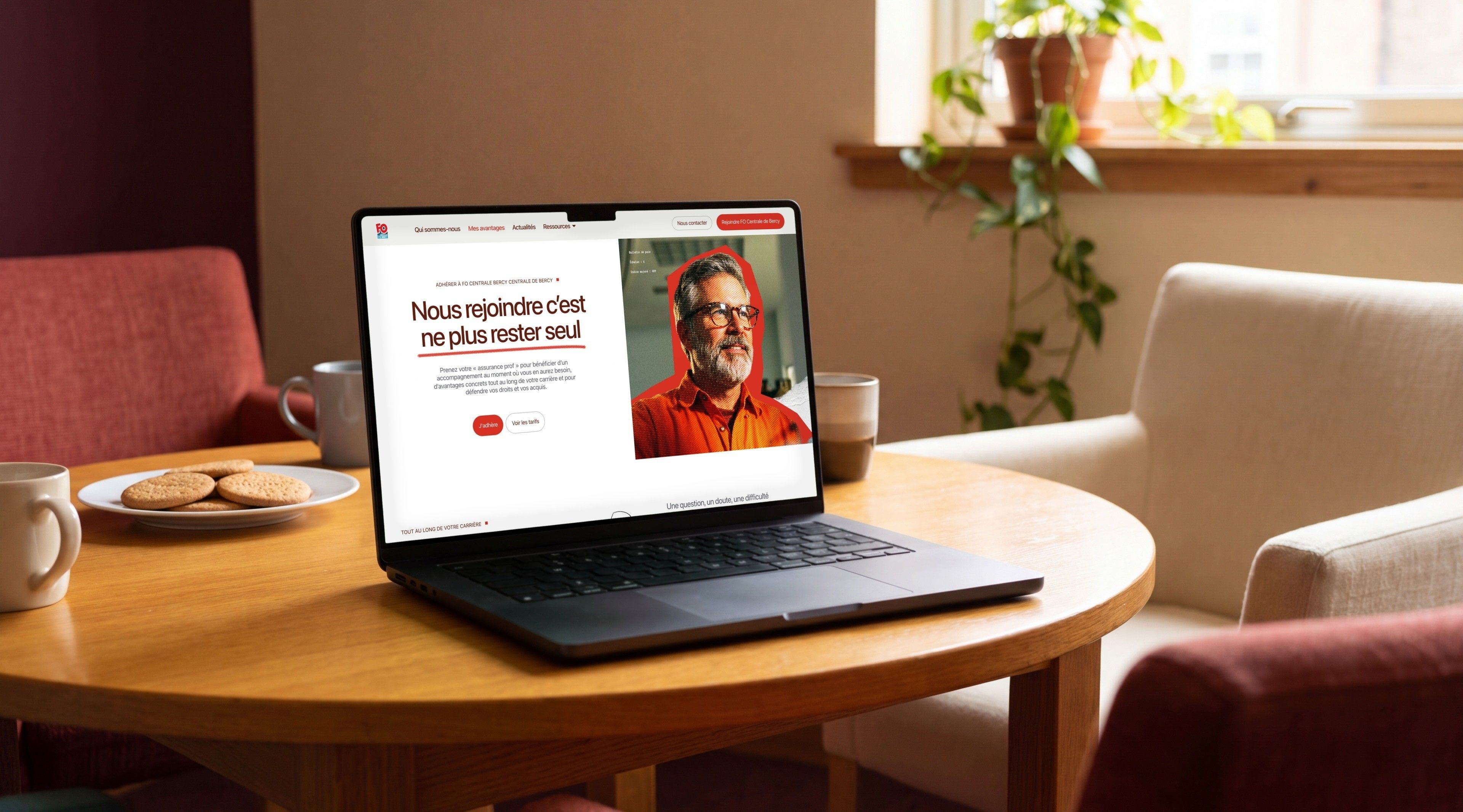 MacBook Pro open on the page “Joining us means no longer being alone”, on a round table with biscuits and coffee