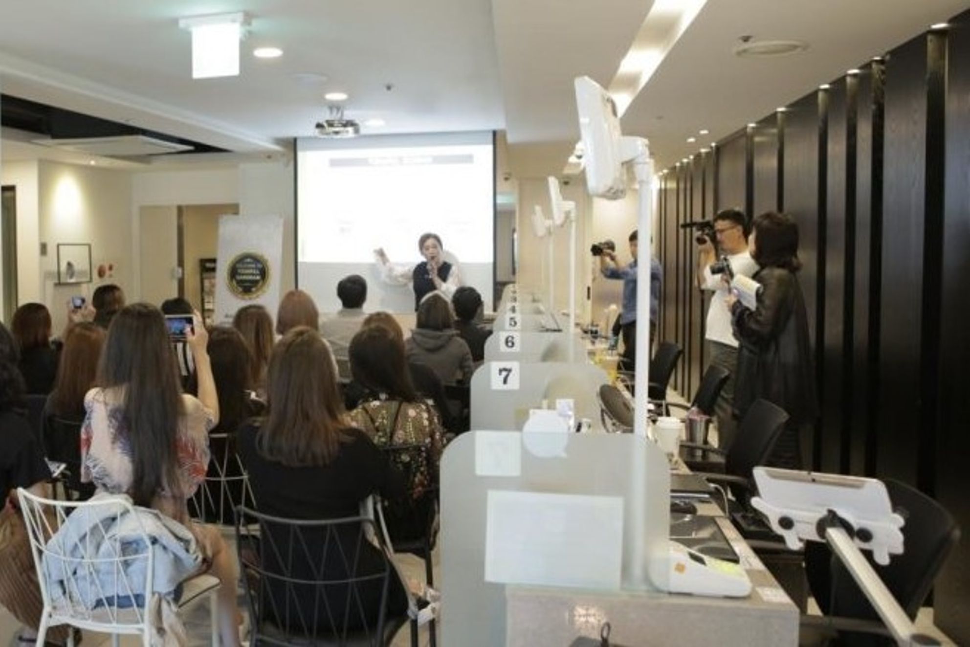 화면 앞에서 마이크를 들고 발표하는 사람과 의자에 앉은 청중, 오른쪽 뒤편에서 촬영하는 취재진
