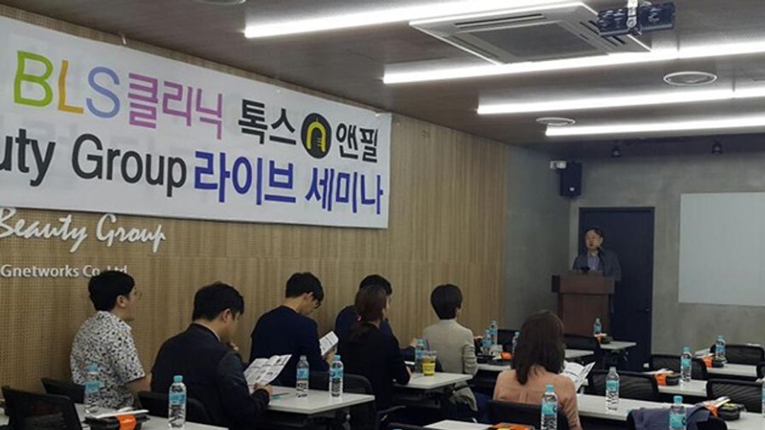 라이브 세미나 현수막이 걸린 강의실에서 참석자들이 책상에 앉아 인쇄물을 보고, 앞쪽 연단에 한 사람이 서 있는 모습