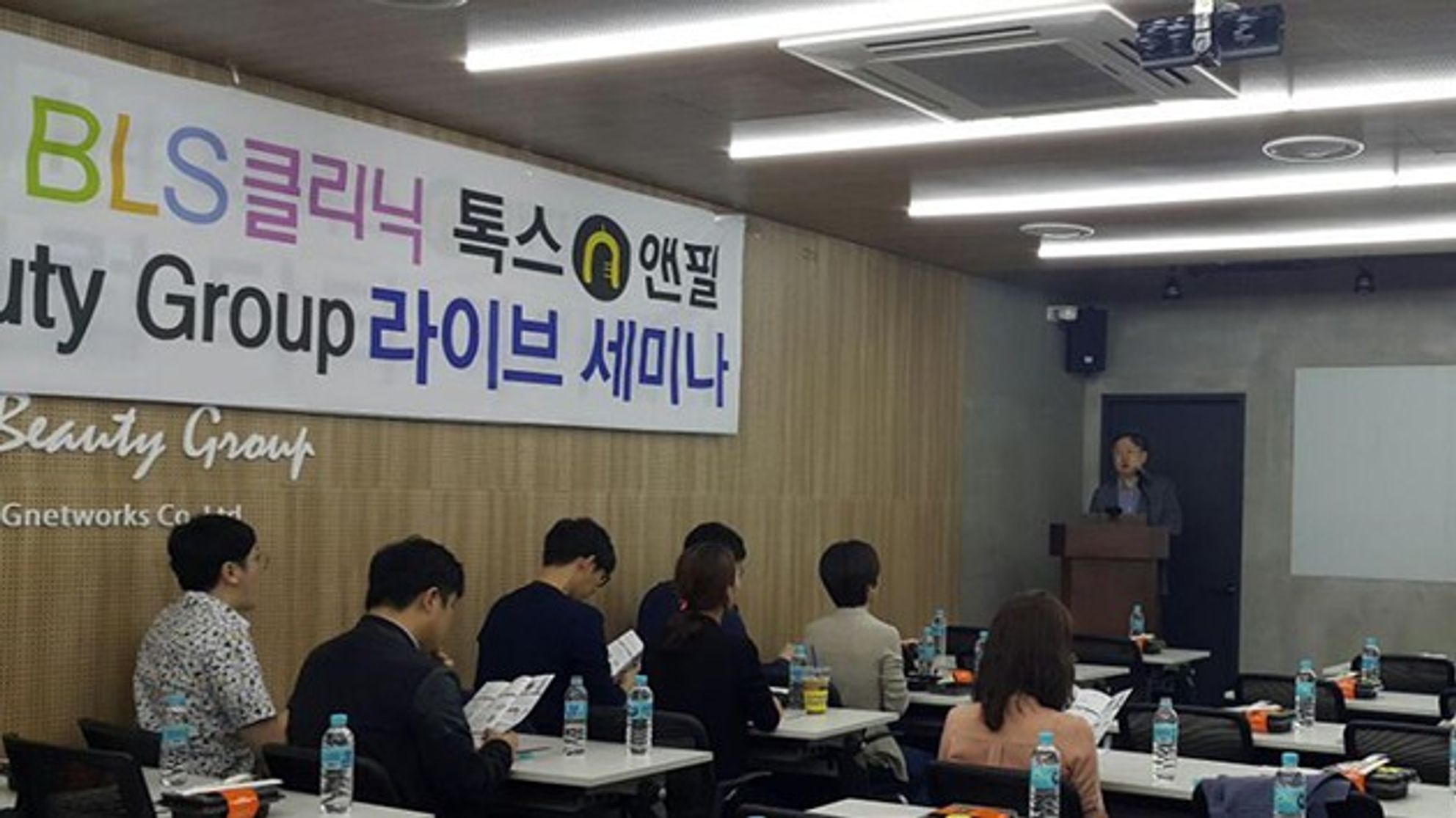 라이브 세미나 현수막이 걸린 강의실에서 참석자들이 책상에 앉아 인쇄물을 보고, 앞쪽 연단에 한 사람이 서 있는 모습