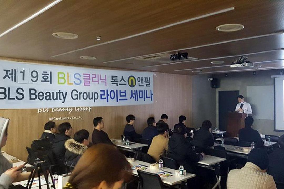 현수막이 걸린 세미나실에서 참석자들이 책상에 앉아 있고 앞쪽 연단에 흰 가운을 입은 사람이 서 있는 모습