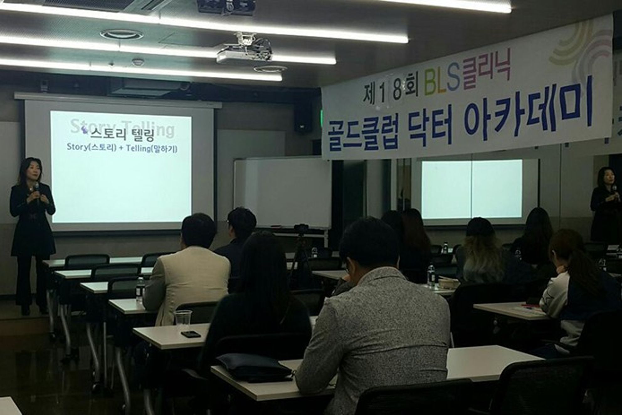 BLS클리닉 현수막이 걸린 강의장에서 참석자들이 앞쪽 스크린과 그 옆에 마이크를 들고 선 사람을 바라보는 뒷모습