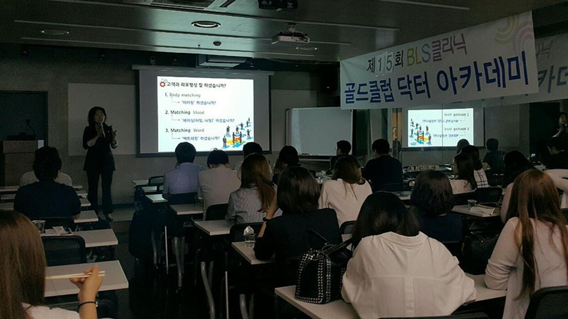 골드클럽 닥터 아카데미 현수막이 걸린 강의실에서 스크린 왼쪽에 선 사람이 마이크를 들고 있고 참석자들이 책상에 앉아 앞을 보는 모습