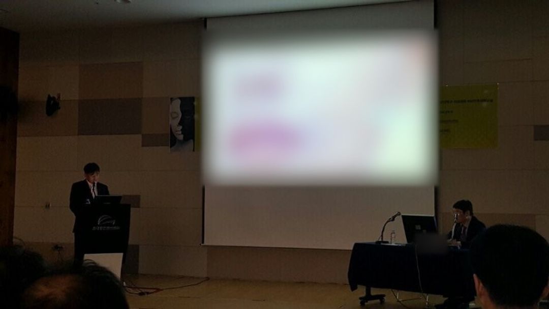 강연장 왼쪽 강연대에 한 사람이 서 있고 큰 화면과 오른쪽 테이블에 앉은 한 사람이 보이는 모습(화면 내용과 명패는 흐리게 처리)