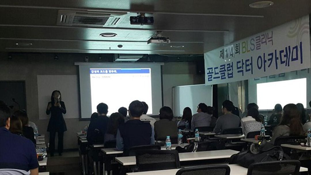 골드클럽 닥터 아카데미 현수막이 걸린 강의장에서 스크린 옆에 한 사람이 마이크를 들고 서 있고 참석자들이 책상에 앉아 앞쪽을 바라보는 모습
