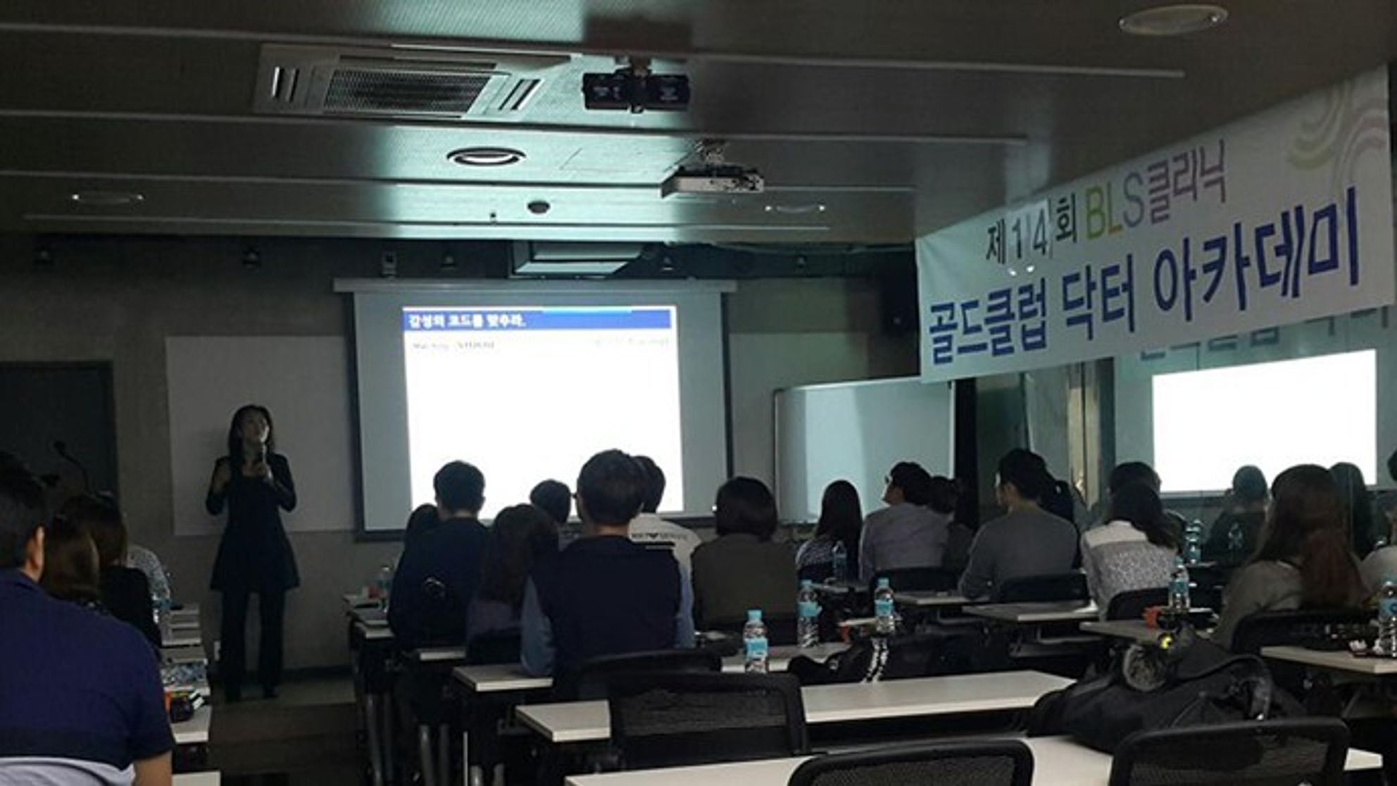 골드클럽 닥터 아카데미 현수막이 걸린 강의장에서 스크린 옆에 한 사람이 마이크를 들고 서 있고 참석자들이 책상에 앉아 앞쪽을 바라보는 모습