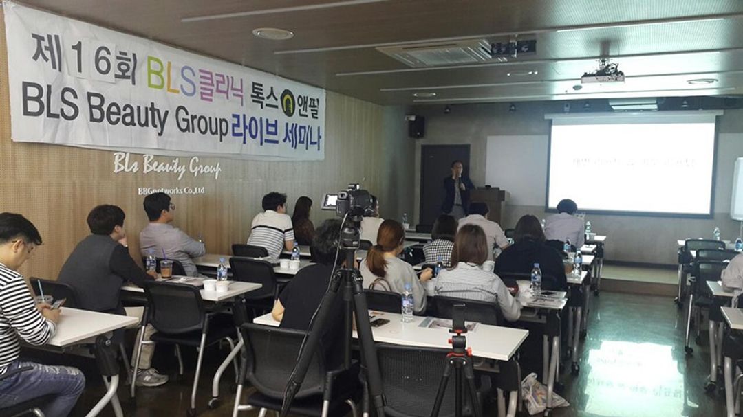 라이브 세미나 현수막이 걸린 강의실에서 참석자들이 책상에 앉아 있고, 앞쪽 연단에서 한 사람이 마이크를 들고 말하는 모습