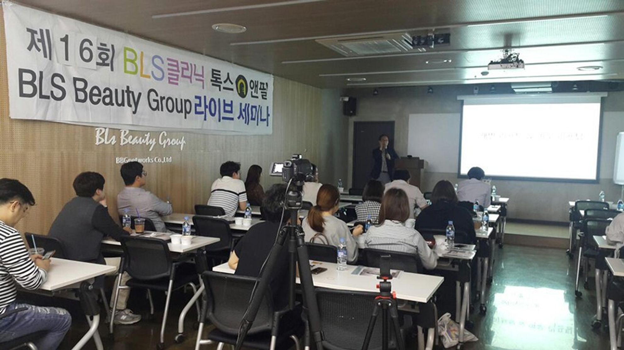 라이브 세미나 현수막이 걸린 강의실에서 참석자들이 책상에 앉아 있고, 앞쪽 연단에서 한 사람이 마이크를 들고 말하는 모습