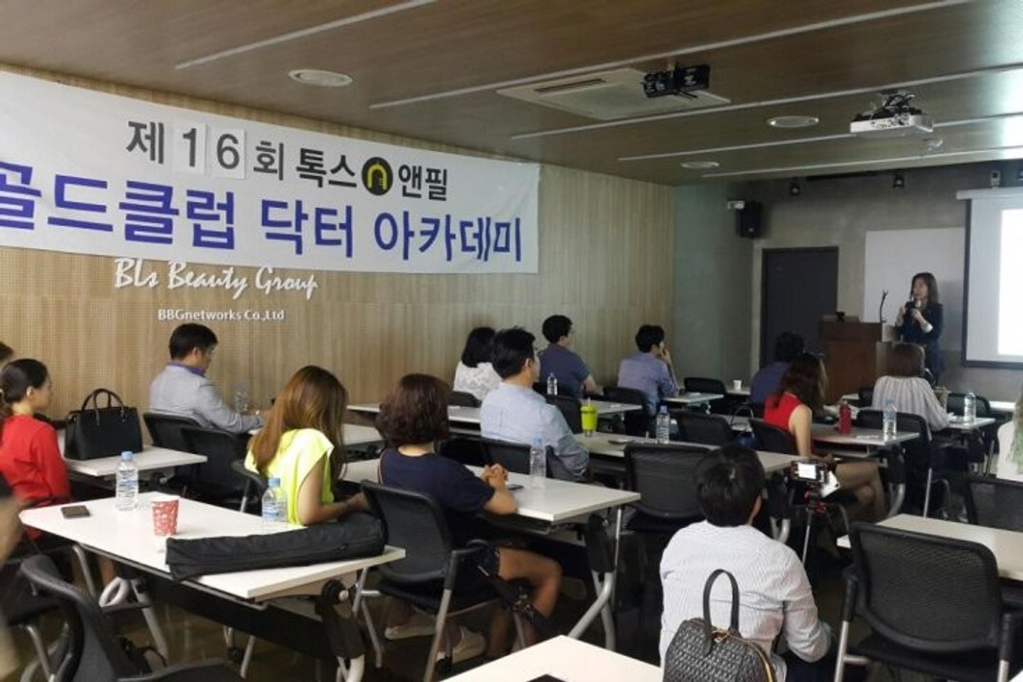 톡스앤필 현수막이 걸린 나무 벽 옆으로 참석자들이 책상에 앉아 있고 오른쪽 앞 연단 옆에 한 사람이 마이크를 들고 서 있는 모습