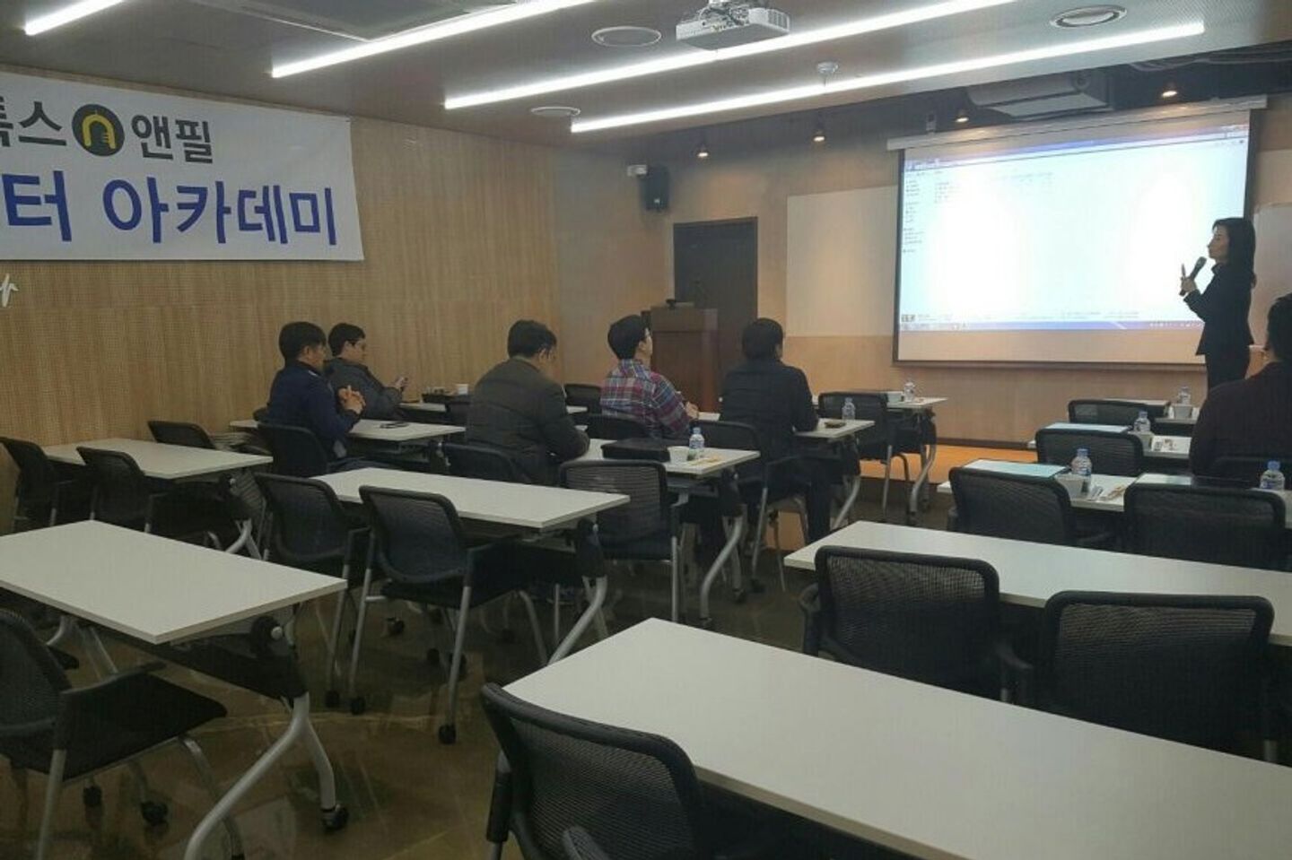 강의장 앞쪽 화면 옆에서 한 사람이 마이크를 들고 말하고, 참석자들이 책상에 앉아 화면을 보는 모습
