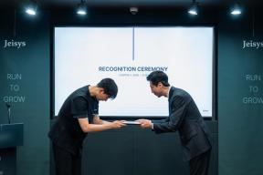 'Jeisys'와 'RUN TO GROW' 글자가 있는 무대 벽 앞, 'RECOGNITION CEREMONY' 화면 아래에서 두 사람이 고개 숙여 인사하며 증서를 주고받고 있다