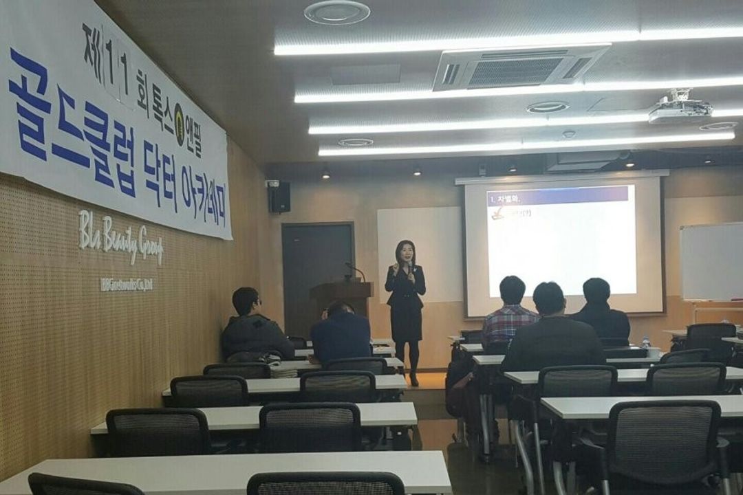 '제11회 톡스앤필 골드클럽 닥터 아카데미' 현수막이 걸린 강의장에서 한 사람이 화면 앞에 서서 마이크를 들고 말하고, 참석자들이 책상에 앉아 듣는 모습