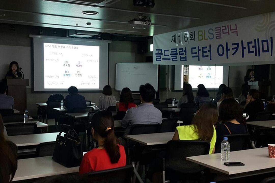 BLS클리닉 현수막이 걸린 강의장에서 참석자들이 책상에 앉아 앞쪽 스크린과 연단에 선 사람을 바라보는 뒷모습