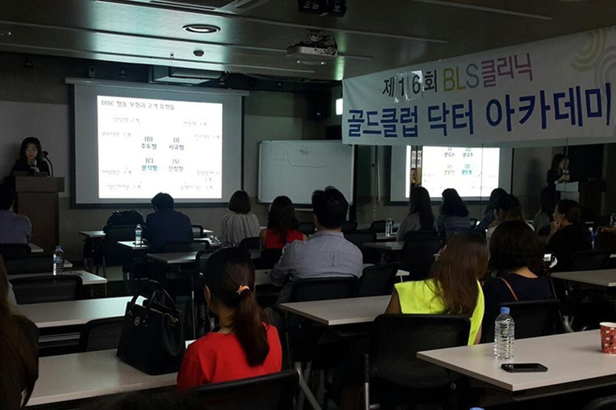 BLS클리닉 현수막이 걸린 강의장에서 참석자들이 책상에 앉아 앞쪽 스크린과 연단에 선 사람을 바라보는 뒷모습