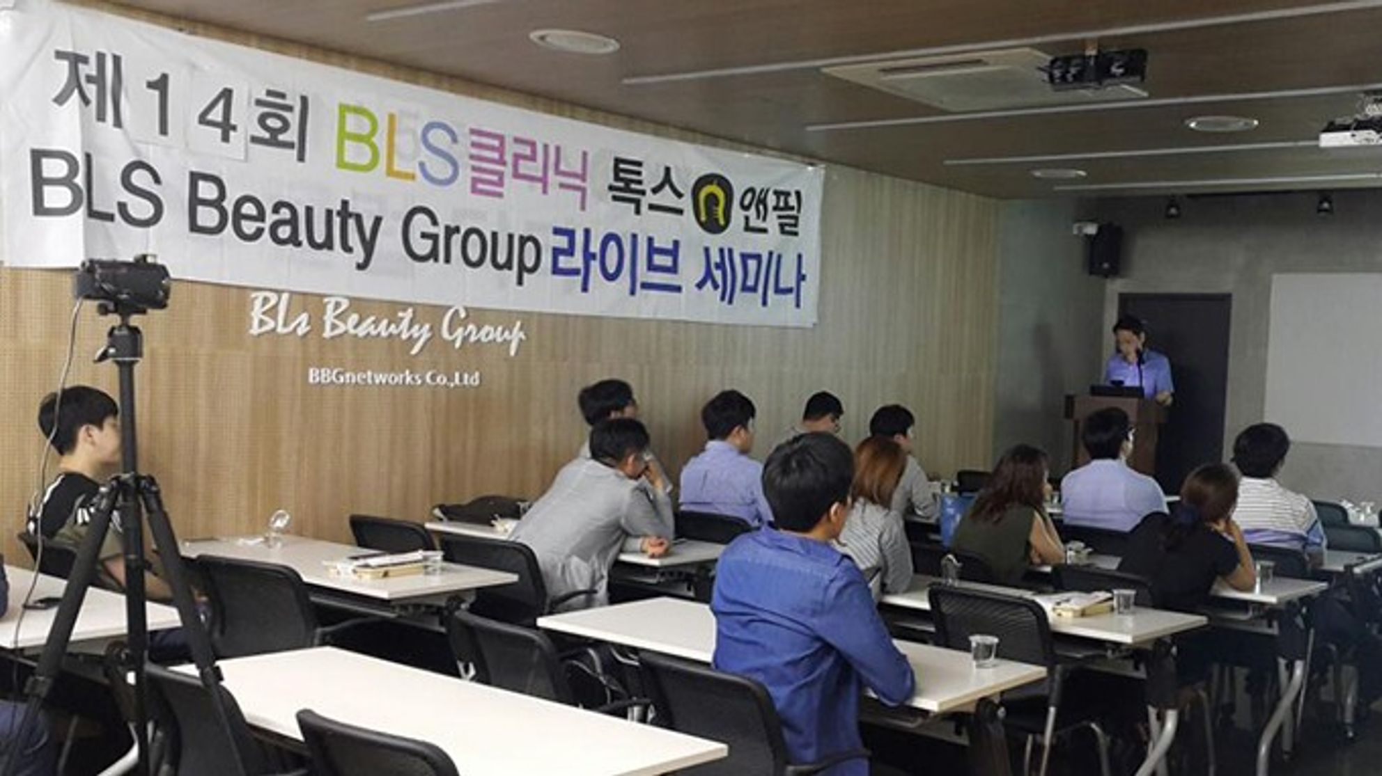 라이브 세미나 현수막이 걸린 강의실에서 참석자들이 책상 앞에 앉아 있고, 앞쪽 연단에 한 사람이 서 있는 모습