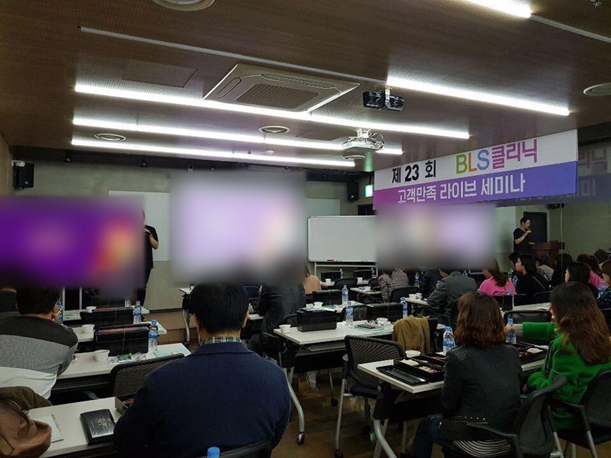 '제23회 BLS클리닉 고객만족 라이브 세미나' 현수막이 걸린 강의실에서 참석자들이 책상에 앉아 앞쪽을 보고 있고 앞에 두 사람이 서 있는 모습(화면 내용은 흐리게 처리)