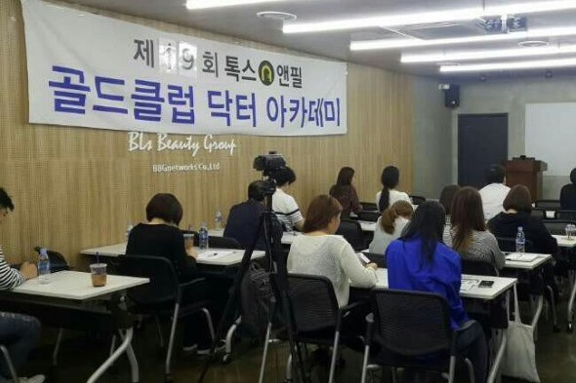 톡스앤필 골드클럽 닥터 아카데미 현수막이 걸린 나무 벽 아래로 참석자들이 책상에 앉아 앞쪽을 바라보는 모습
