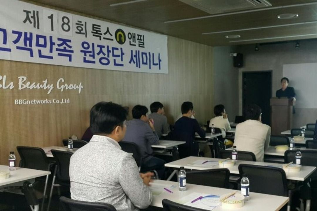 톡스앤필 고객만족 원장단 세미나 현수막이 걸린 강의장에서 한 사람이 단상에서 발표하고 참석자들이 책상에 앉아 듣는 모습