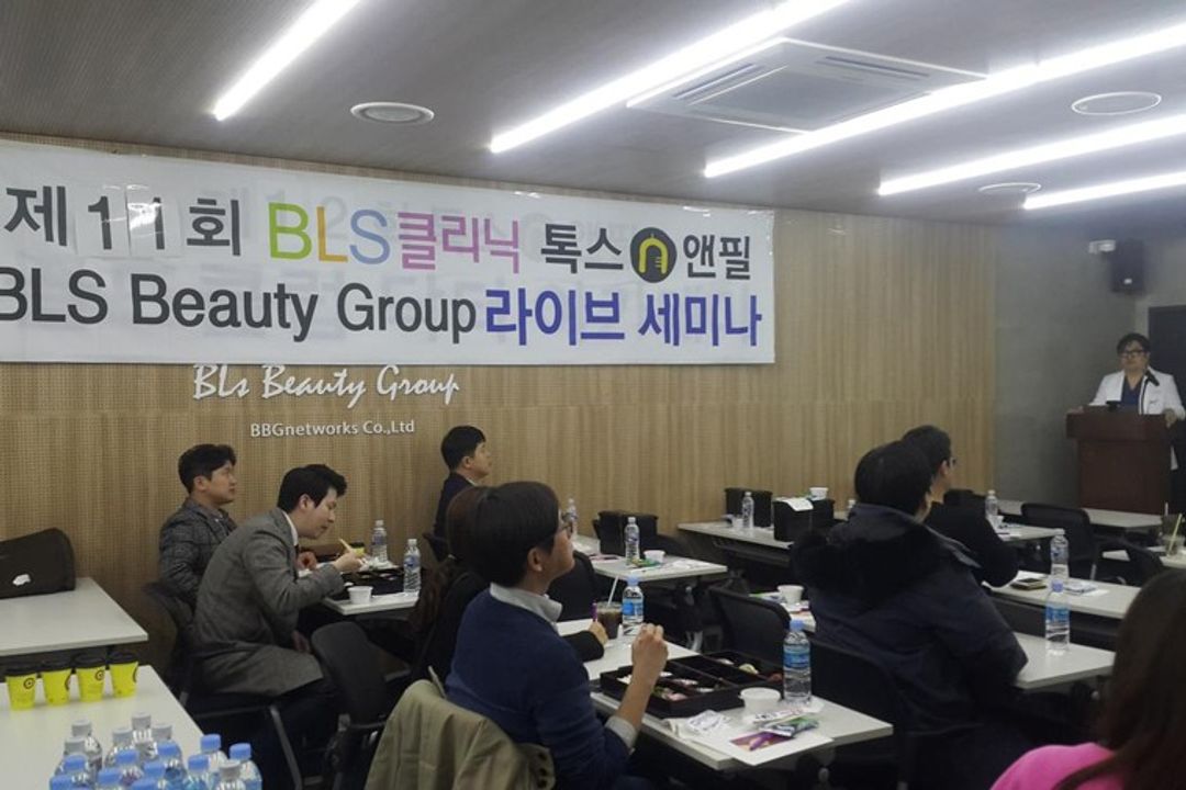 현수막이 걸린 세미나실에서 참석자들이 책상에 앉아 있고 앞쪽 연단에 흰 가운을 입은 사람이 서 있는 모습