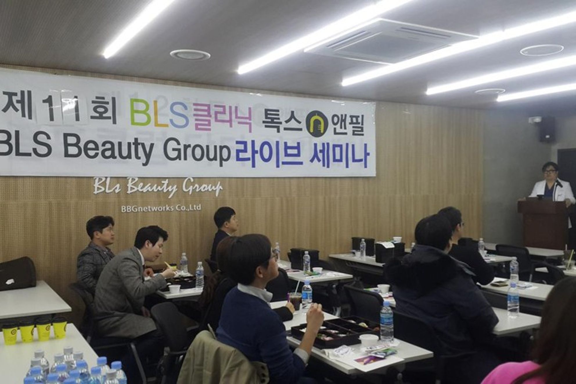 현수막이 걸린 세미나실에서 참석자들이 책상에 앉아 있고 앞쪽 연단에 흰 가운을 입은 사람이 서 있는 모습