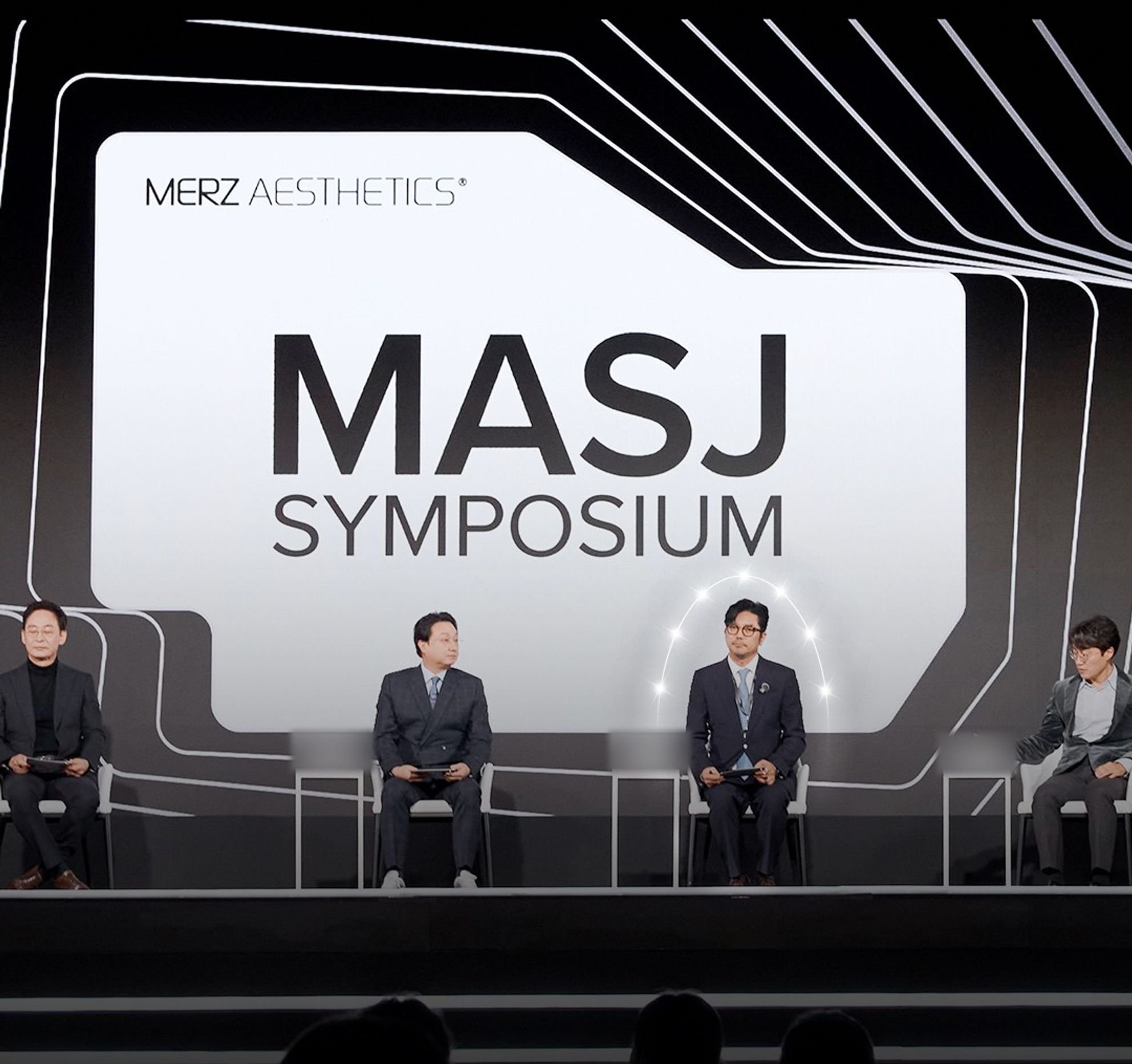 'MASJ SYMPOSIUM' 대형 화면 아래 무대에 네 사람이 의자에 앉아 있는 모습(명패 글자는 흐리게 처리)