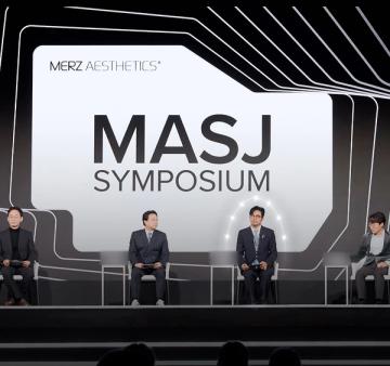 'MASJ SYMPOSIUM' 대형 화면 아래 무대에 네 사람이 의자에 앉아 있는 모습(명패 글자는 흐리게 처리)