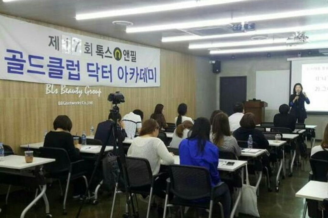 톡스앤필 현수막이 걸린 나무 벽 강의장에서 참석자들이 책상에 앉아 오른쪽 앞 스크린 옆에 마이크를 들고 선 사람을 바라보는 모습