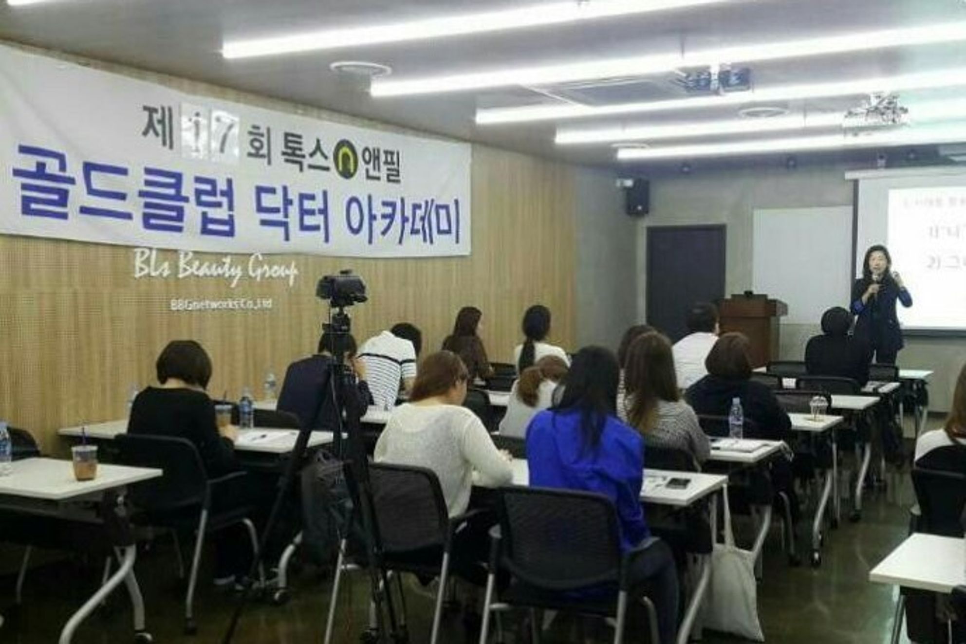 톡스앤필 현수막이 걸린 나무 벽 강의장에서 참석자들이 책상에 앉아 오른쪽 앞 스크린 옆에 마이크를 들고 선 사람을 바라보는 모습