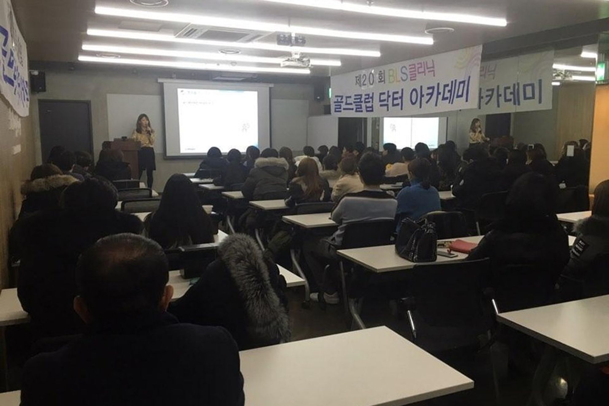BLS클리닉 현수막이 걸린 강의장에서 참석자들이 책상에 앉아 앞쪽 스크린 옆에서 마이크를 든 사람을 바라보는 뒷모습