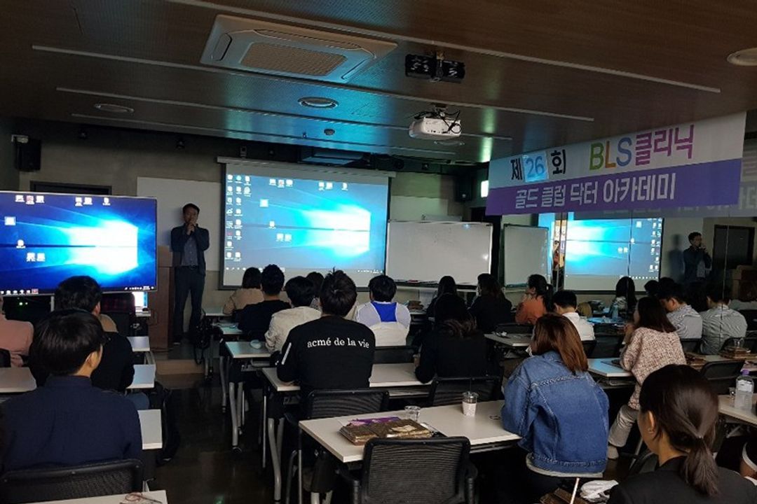 골드 클럽 닥터 아카데미 현수막이 걸린 강의장에서 한 사람이 앞쪽에 서서 마이크를 들고 말하고, 참석자들이 책상에 앉아 듣는 모습