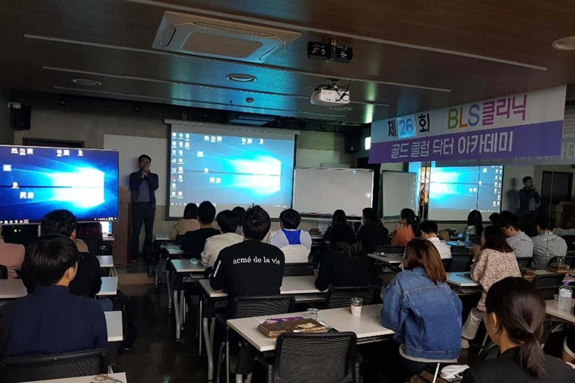 골드 클럽 닥터 아카데미 현수막이 걸린 강의장에서 한 사람이 앞쪽에 서서 마이크를 들고 말하고, 참석자들이 책상에 앉아 듣는 모습