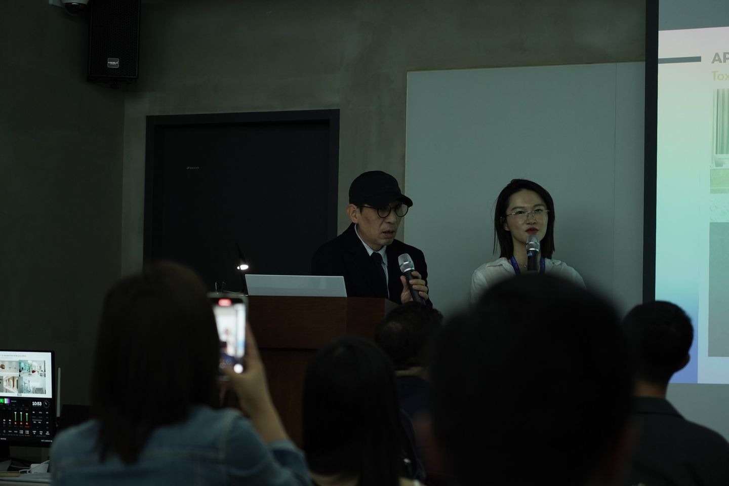 어두운 강의실 연단에서 발표자가 마이크로 설명하고 옆에 선 사람도 마이크를 들고 있으며, 사진 앞쪽 객석의 참석자가 휴대전화를 들어 촬영하고 있다