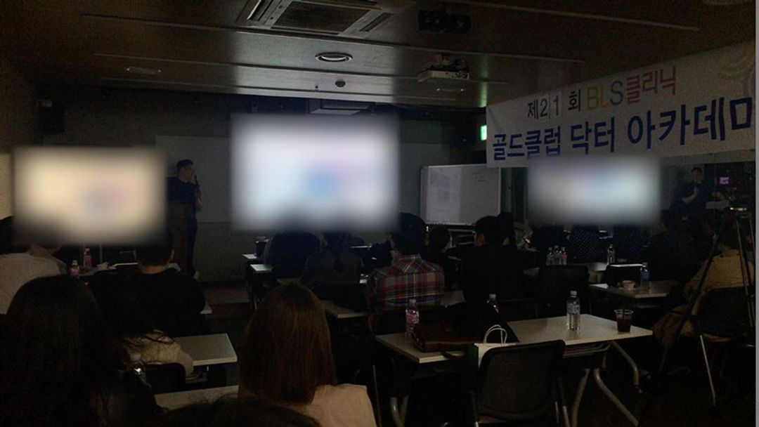 '제21회 BLS클리닉 골드클럽 닥터 아카데미' 현수막이 걸린 어두운 강의실에서 참석자들이 앉아 앞쪽 화면 쪽을 보고 있고 앞에 한 사람이 서 있는 모습(화면 내용은 흐리게 처리)