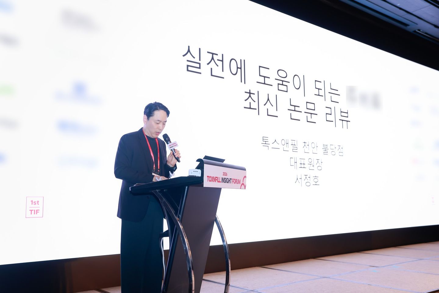 '2026 TOXNFILL INSIGHT FORUM' 연단에서 연자가 마이크를 들고 발표하고 있고, 뒤 화면에 '최신 논문 리뷰'와 '톡스앤필 천안 불당점 대표원장 서정호' 글자가 보인다